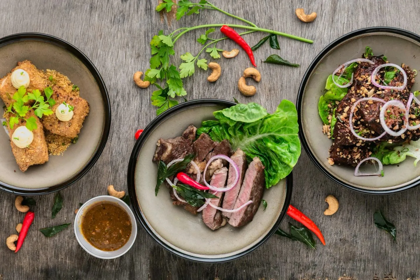 Overhead view of several grey ceramic plates with sliced meat, greens and small bowls of sauce